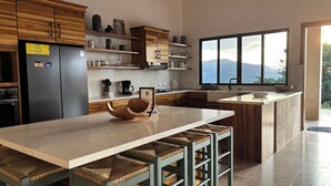 A modern kitchen with wooden cabinets, a large island with bar stools, a refrigerator, and a window with a mountain view. - Panoramic Lake Arenal Sunset View Villa with AC (Nuevo Arenal)