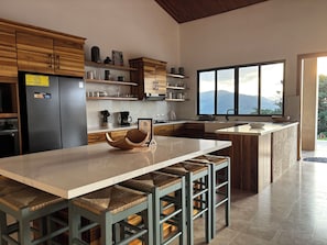 A modern kitchen with wooden cabinets, a large island with bar stools, a refrigerator, and a window with a mountain view.