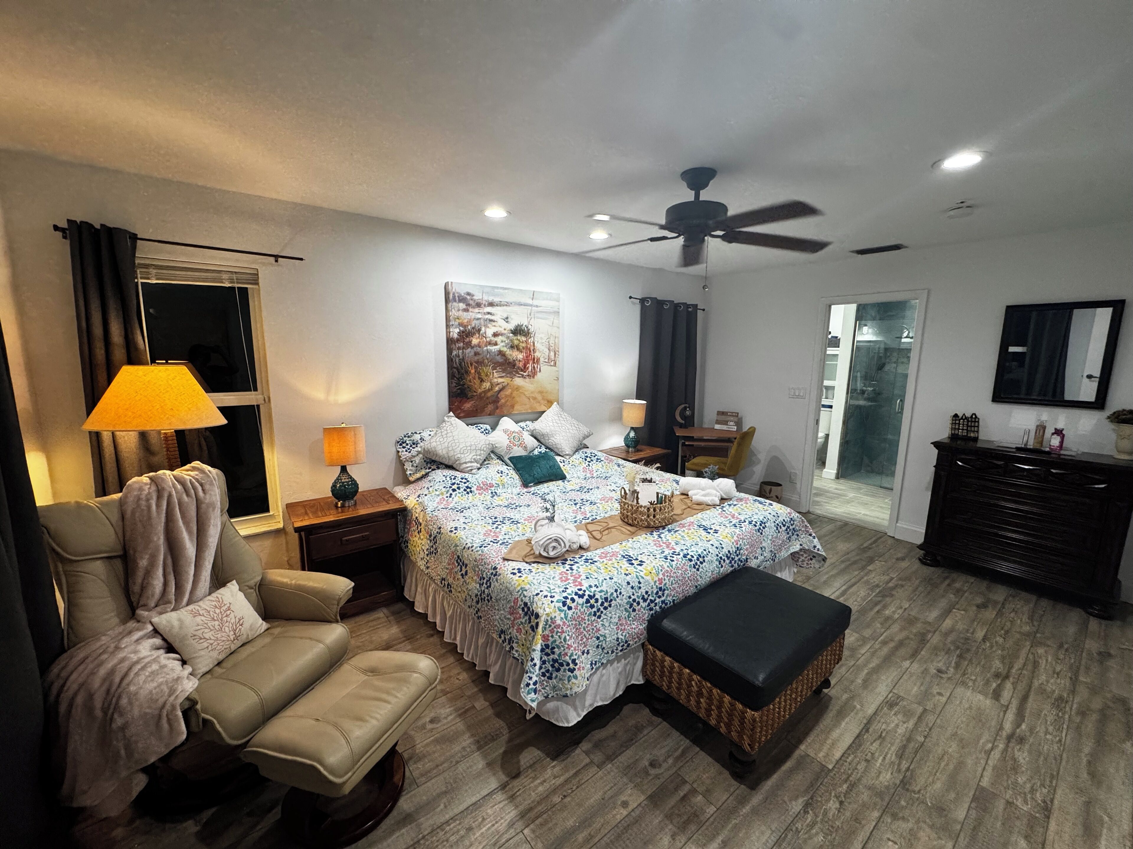 A bedroom with a bed, a chair, a desk, a dresser, a ceiling fan, and a bathroom with a glass door.
