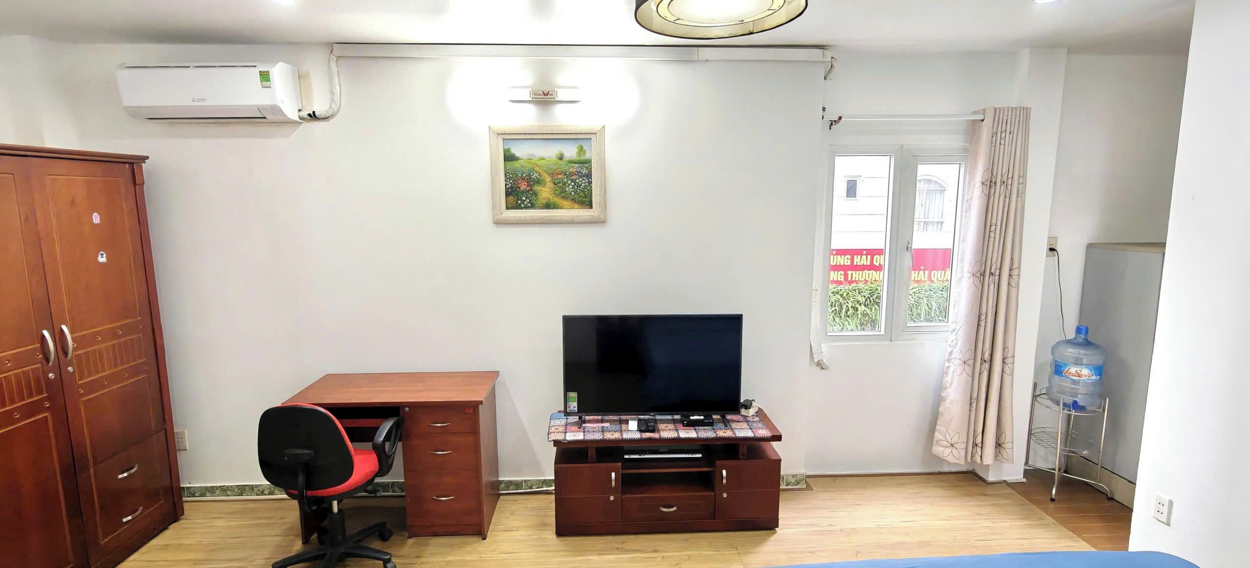 A bedroom with a wooden desk, a television on a stand, a wardrobe, and a window with curtains.