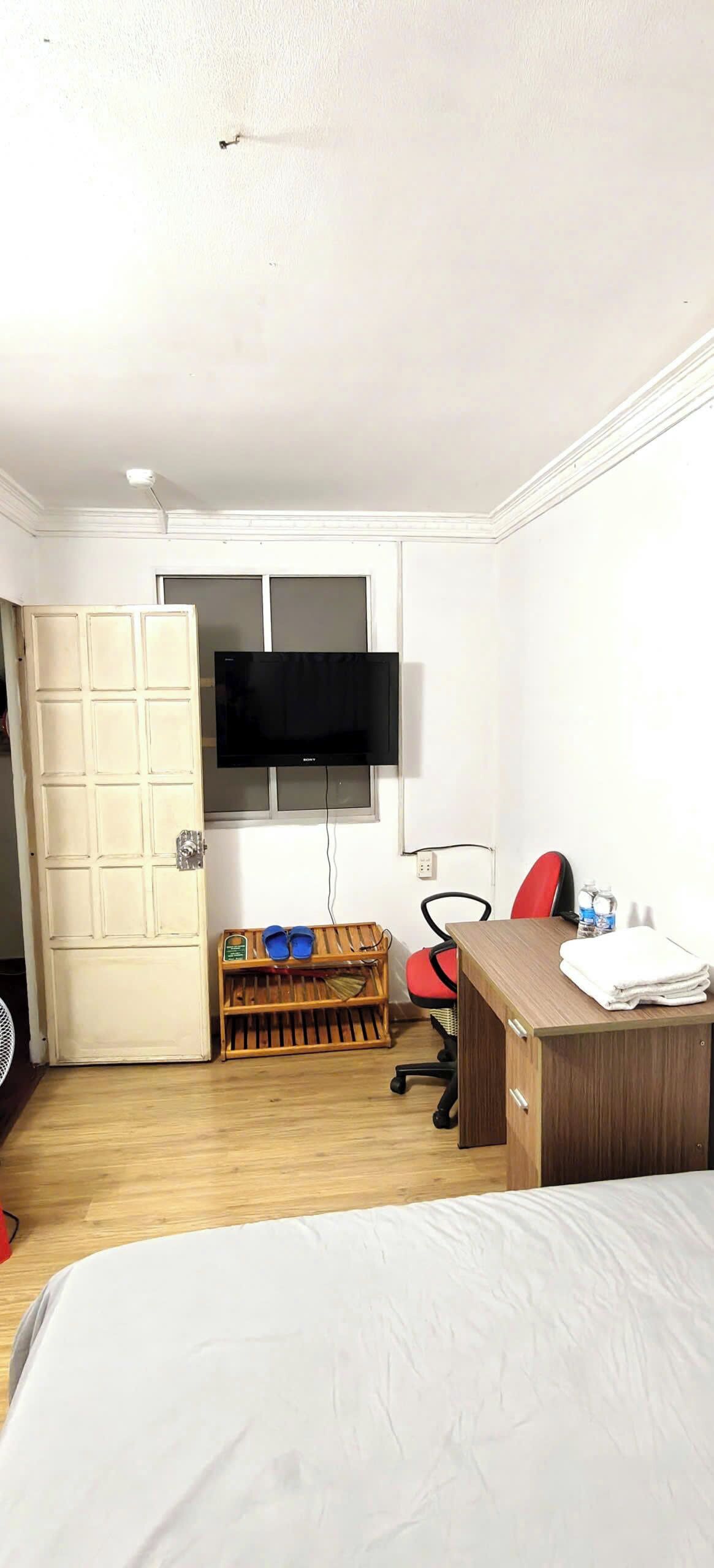 A hotel room with a bed, a desk with a chair, a television mounted on the wall, a wooden bench with slippers, and a door.