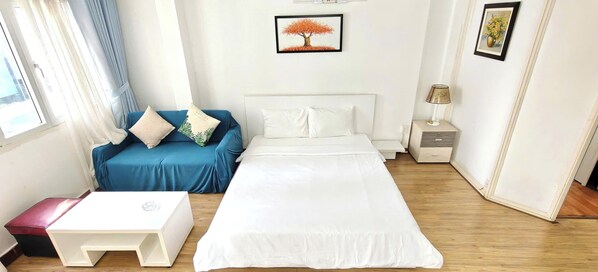 A hotel room with a bed, a blue sofa, a small white table, and a nightstand with a lamp. - Lunaria Maison Japan Town Kitchen Suite (Ho Chi Minh City)