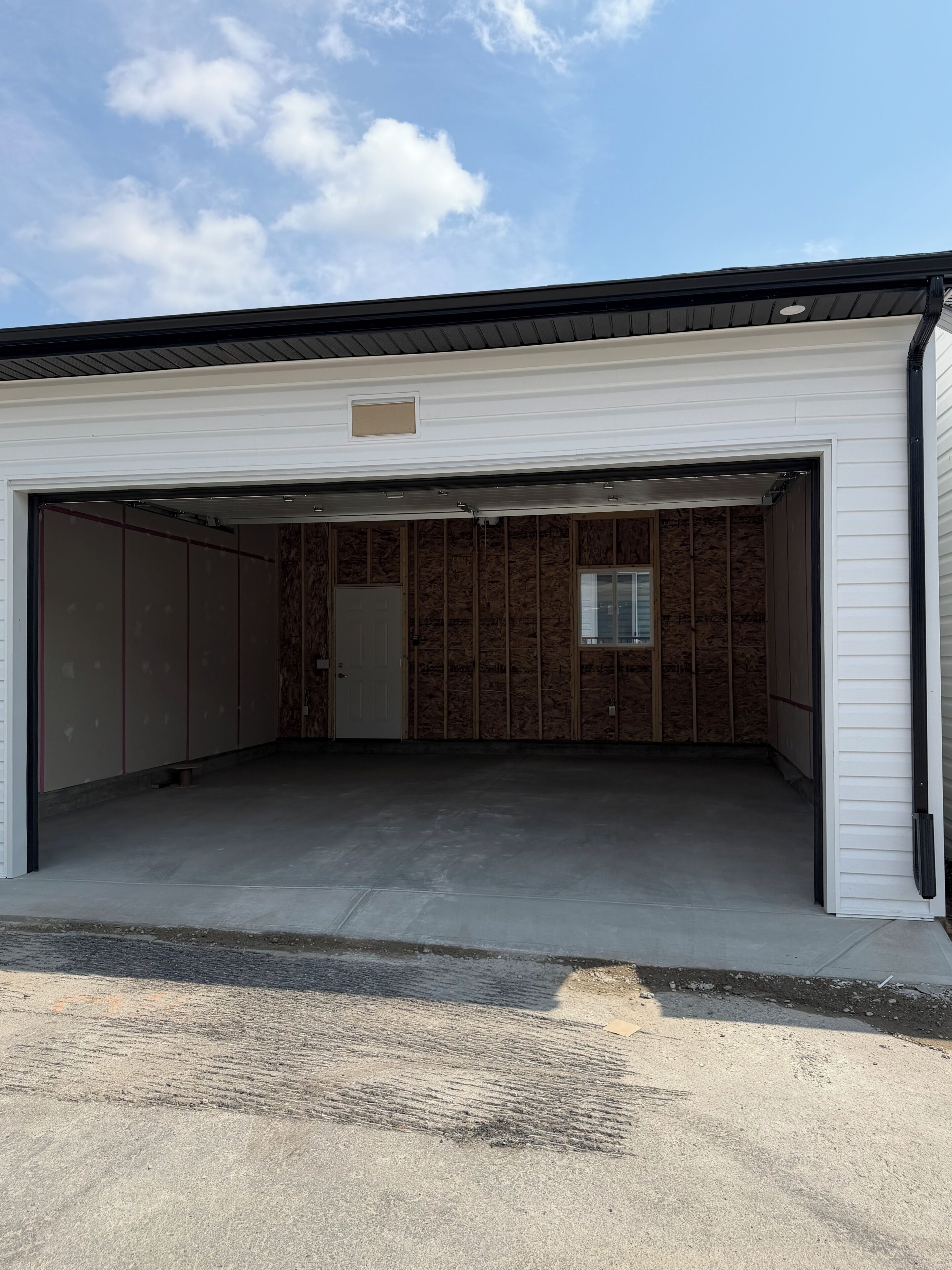Un bâtiment avec une porte de garage ouverte, révélant un intérieur inachevé avec une structure en bois et une petite fenêtre.