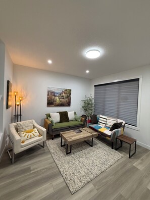 A modern living room with a sofa, armchair, coffee table, and a large window with blinds. - Slice of Heaven - Near Spruce Meadows - 2 Car Garage Central AC avail -Sleeps 8  (Calgary)