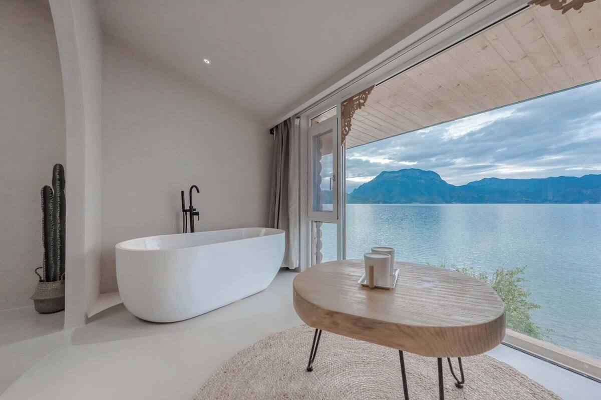 Une salle de bain moderne avec une baignoire sur pieds, une table en bois avec une tasse, et une vue sur un lac et des montagnes.