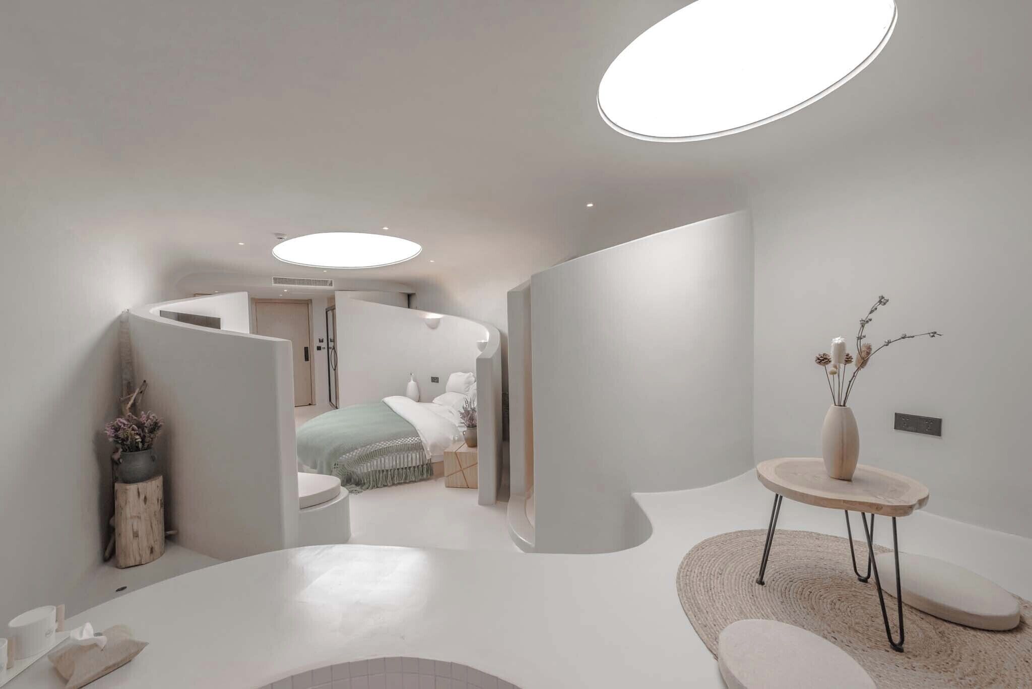 Une chambre d’hôtel moderne et minimaliste, avec un mur blanc incurvé, une table ronde ornée d’un vase et un lit recouvert d’une couverture verte.