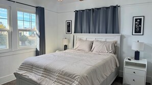 Ein ordentlich bezogenes Bett mit weißem Kopfteil, ein Nachttisch mit Lampe und ein Fenster mit Vorhängen.