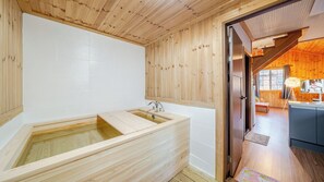 Un intérieur en bois avec une baignoire en bois, une porte en bois donnant sur la cuisine et une vue sur l’extérieur.