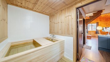 A wooden interior with a wooden bathtub, a wooden door leading to a kitchen, and a view of the outside.