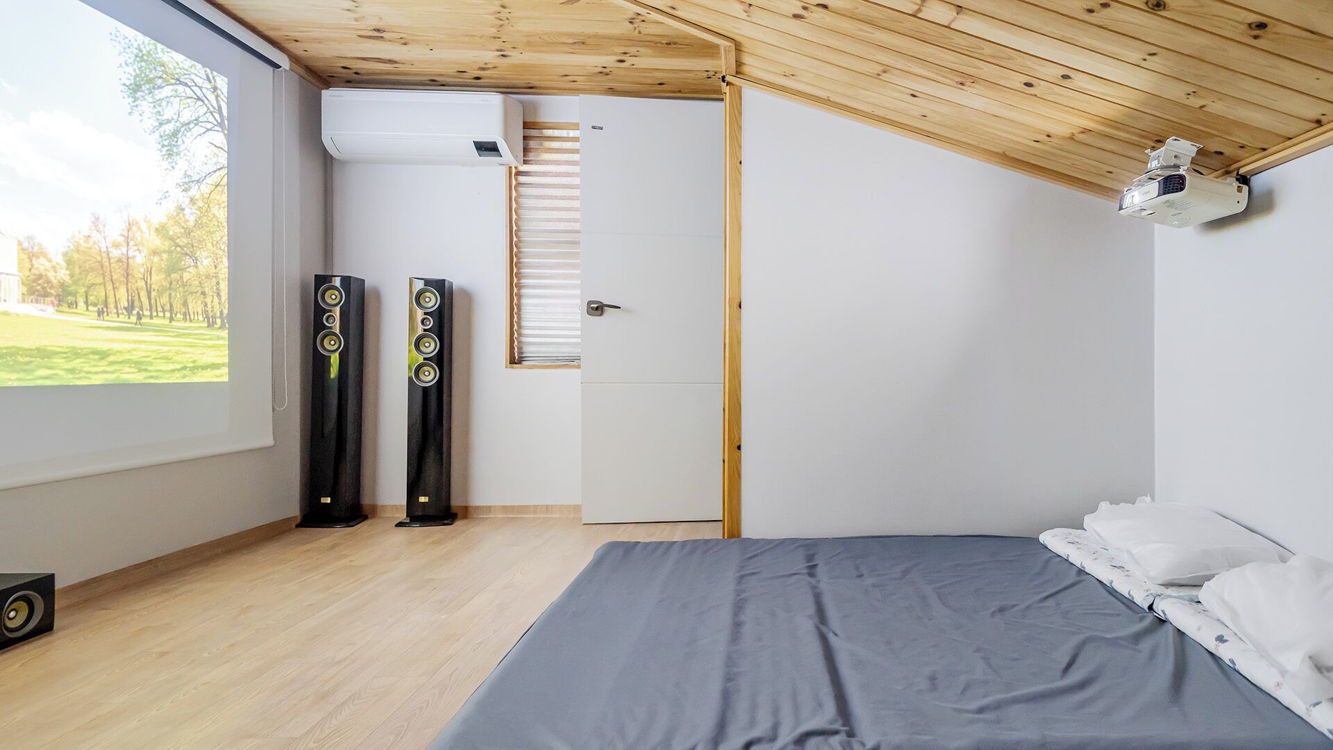 A bedroom with a bed, two speakers, a window with a view of trees, and a door with blinds.