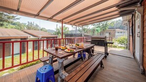 Een overdekt terras buiten met een houten eettafel, een barbecue en uitzicht op de omgeving.