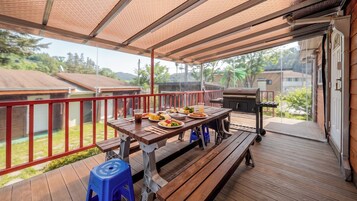 A covered outdoor patio with a wooden table set for dining, a barbecue grill, and a view of the surrounding neighborhood.