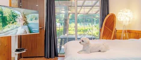 A hotel room with a bed, a television, a wooden wardrobe, a large window with curtains, and a small table.