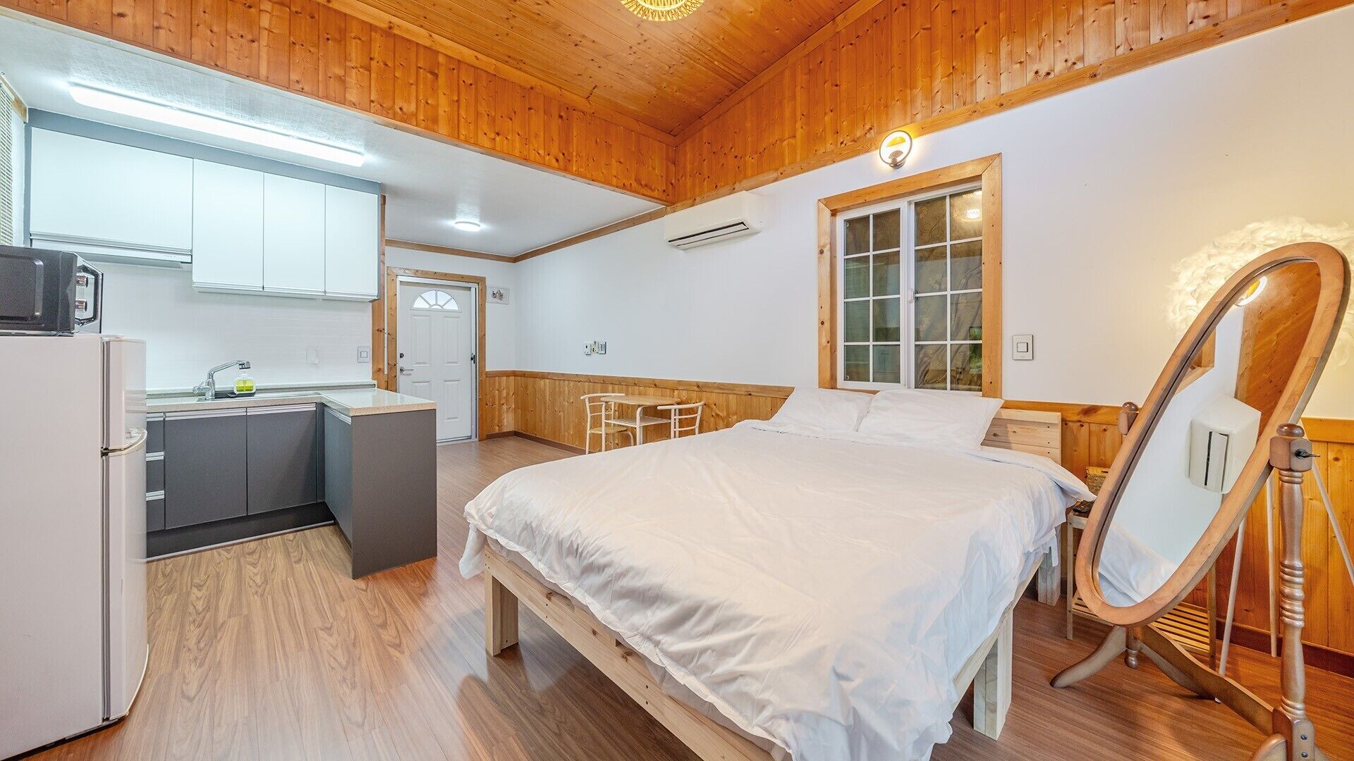 A room with a bed, a desk, a mirror, and a kitchenette.