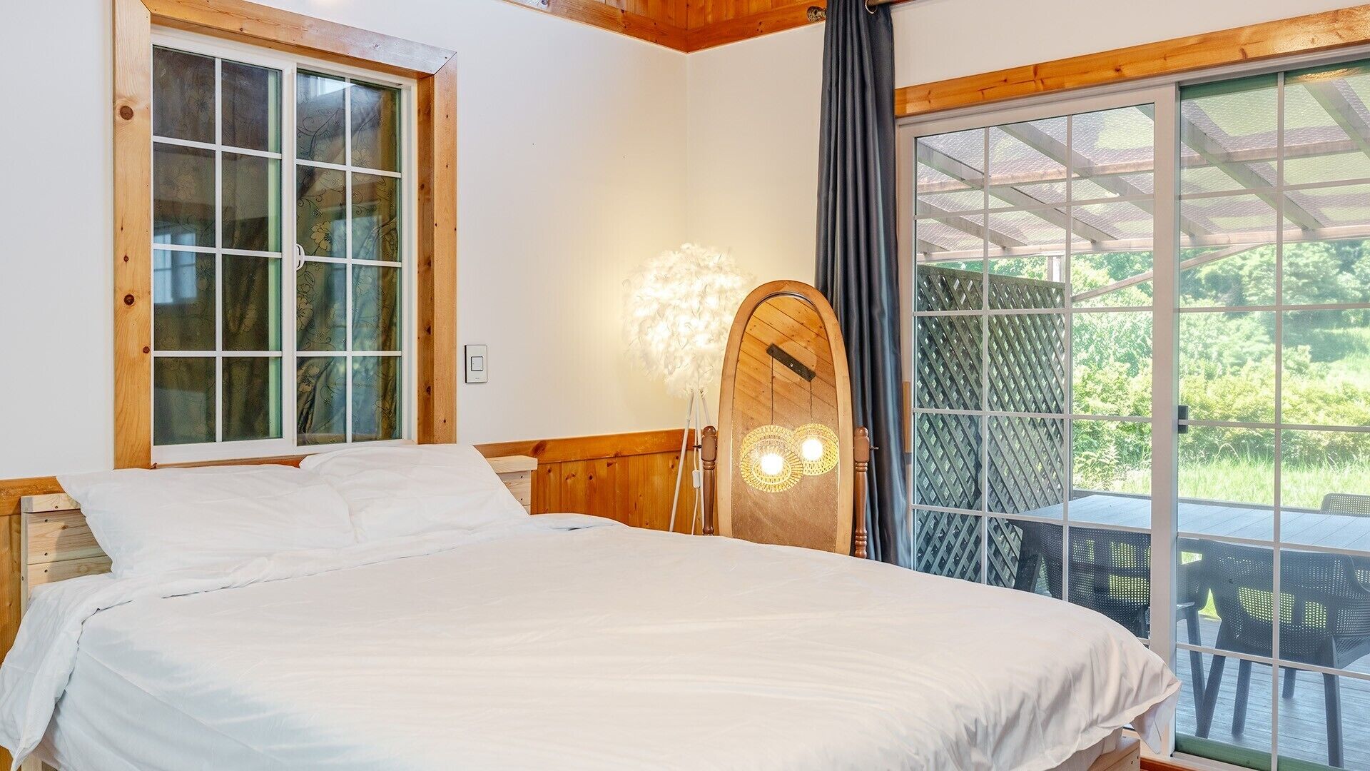 A bedroom with a bed, a window with curtains, a wall-mounted light fixture, and a view of the outdoors.