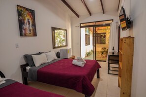 A hotel room with two beds, a painting, a TV, and a sliding glass door to the outside. - Classic Quadruple Room (Monteverde)