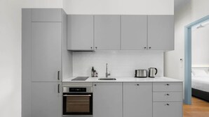 Une cuisine compacte avec des meubles gris, un four encastré, un évier et un plan de travail sur lequel se trouvent une bouilloire et une machine à café.