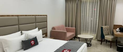 A hotel room with a bed, a pink armchair, a small round table, and a desk with a chair.