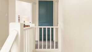 Una escalera conduce a una habitación con una puerta verde azulada, paredes blancas y una pequeña mesa con una lámpara.