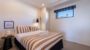 Ein Schlafzimmer mit einem Bett, einem Nachttisch, einer Lampe, einem gerahmten Bild und einem Fenster mit Blick auf einen Turm.