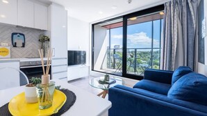 A modern living room with a sofa, a dining table with a yellow placemat, a kitchen area, and a large window with a city view.