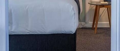Ein ordentlich bezogenes Bett mit weißer Bettwäsche und einem roten Kopfkissen, eine Nachttischlampe und ein kleiner Tisch mit einer Uhr.