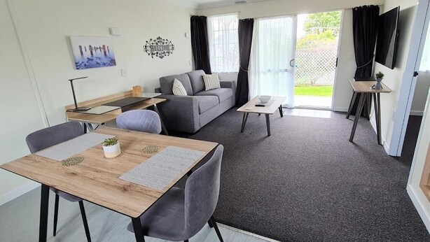A modern living room with a sofa, a dining table, and a TV mounted on the wall. - Kings Court Motel (Whanganui)