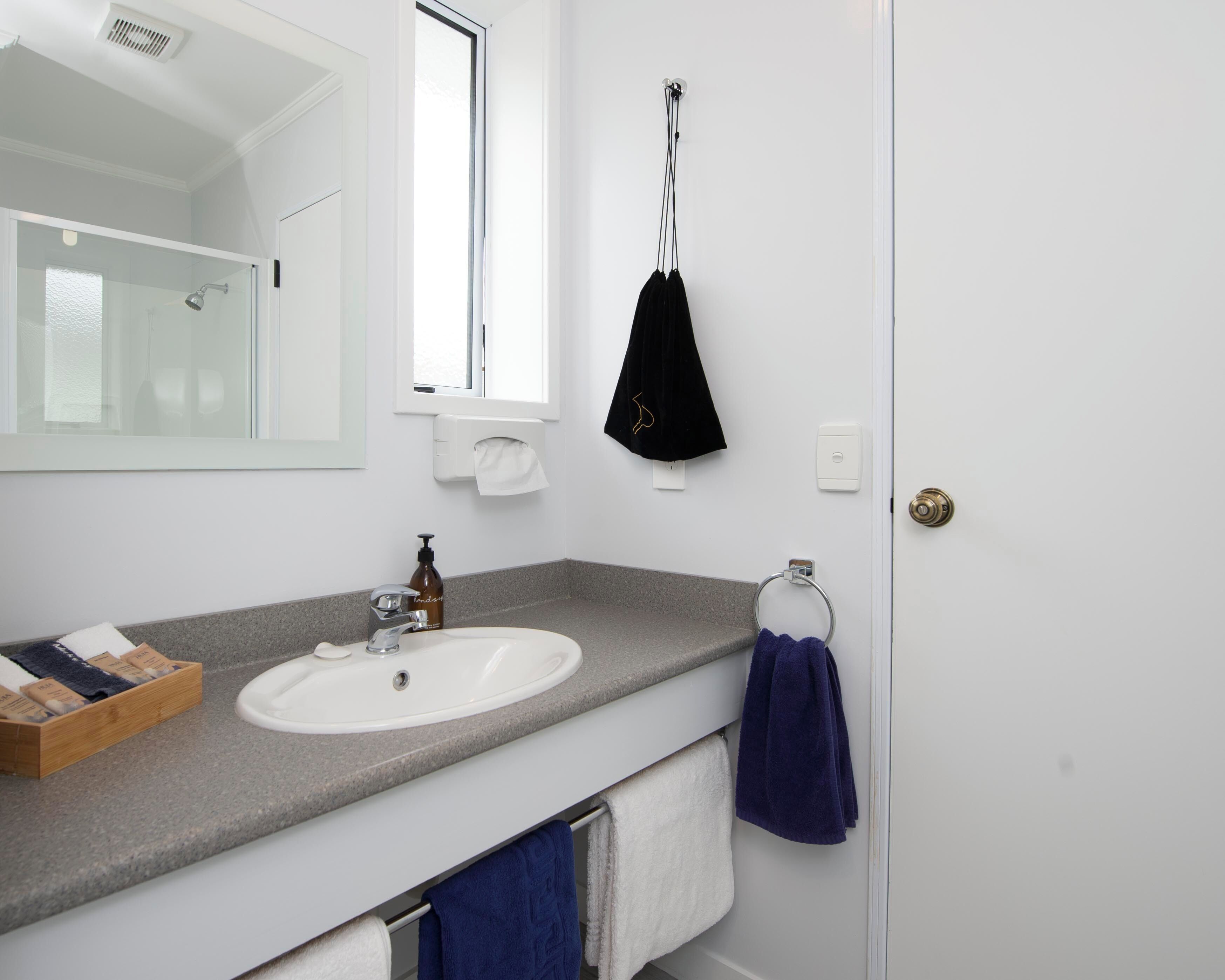 Un baño con un lavamanos blanco, un espejo, una ventana, un perchero para toallas y una puerta.