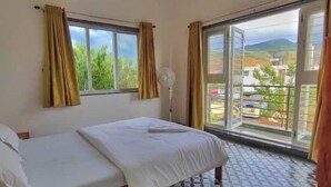 A bedroom with a bed, a window with curtains, a standing fan, and a view of the outside.