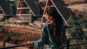 Eine Person steht auf einem Balkon mit Blick auf eine Reihe moderner Gebäude mit dreieckigen Dächern, im Hintergrund eine hügelige Landschaft.