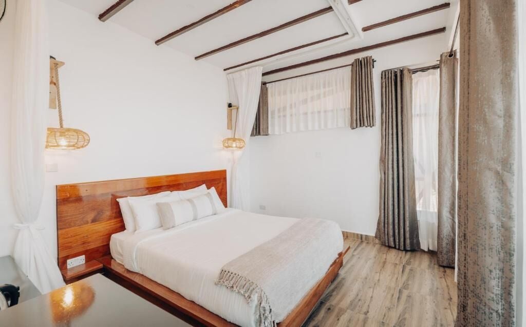 A bedroom with a wooden bed, white bedding, a bedside table, a hanging light, and a window with curtains.