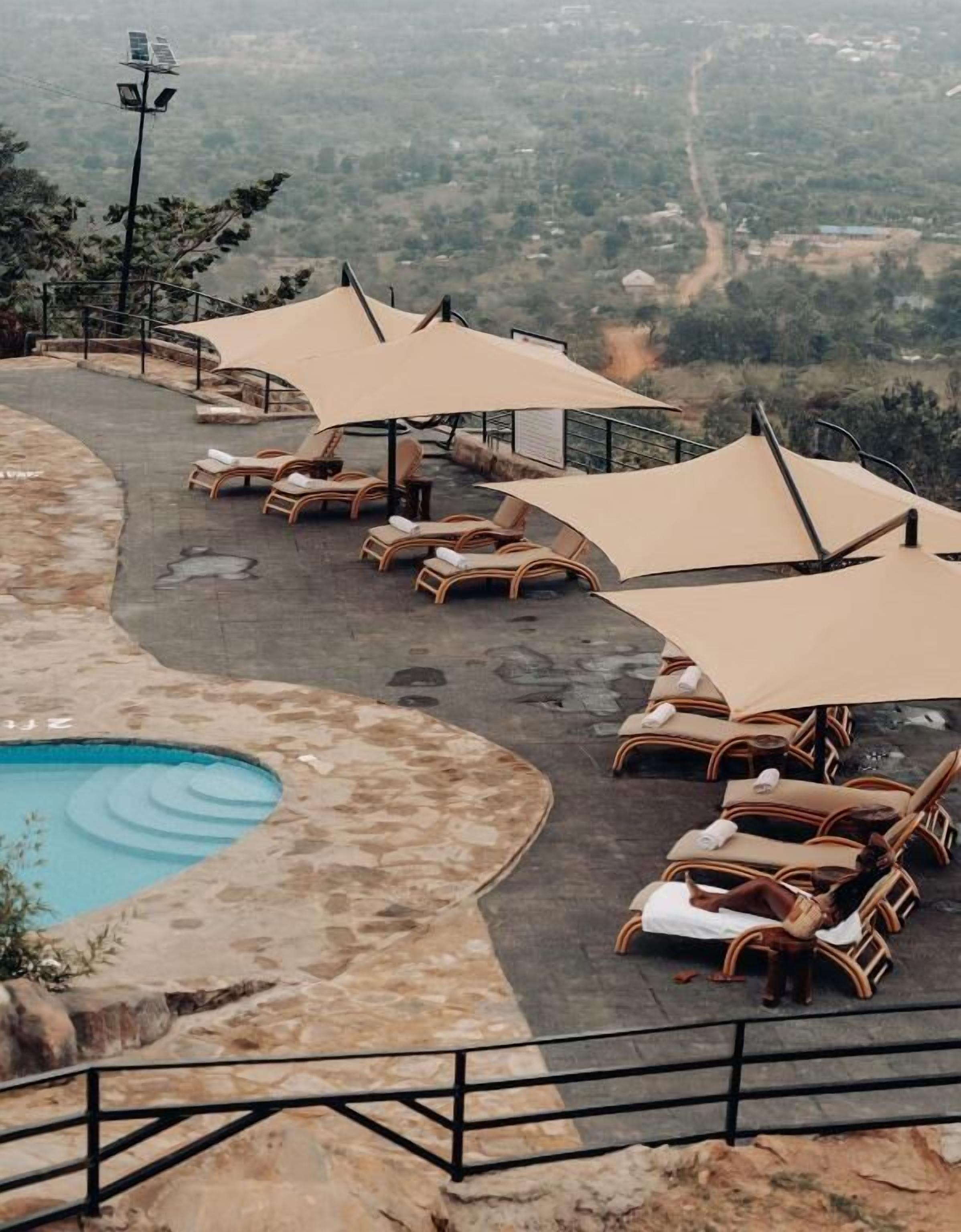 A poolside area with lounge chairs and umbrellas, surrounded by a stone deck and a view of a valley.
