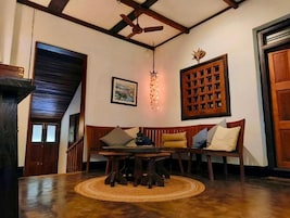 Una habitación con techo de madera, ventilador de techo, banco de madera con cojines, mesa con tapete tejido y una lámpara colgante decorativa.