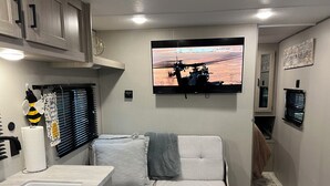 Interior compacto de una autocaravana con kitchenette, sofá, televisor montado y ventana con persianas.