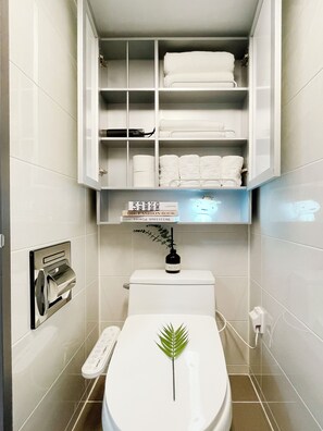 A modern bathroom with a toilet, a shelf with folded towels, a toilet paper holder, and a small plant on the toilet seat. - MZstay Seomyeon (Busan)