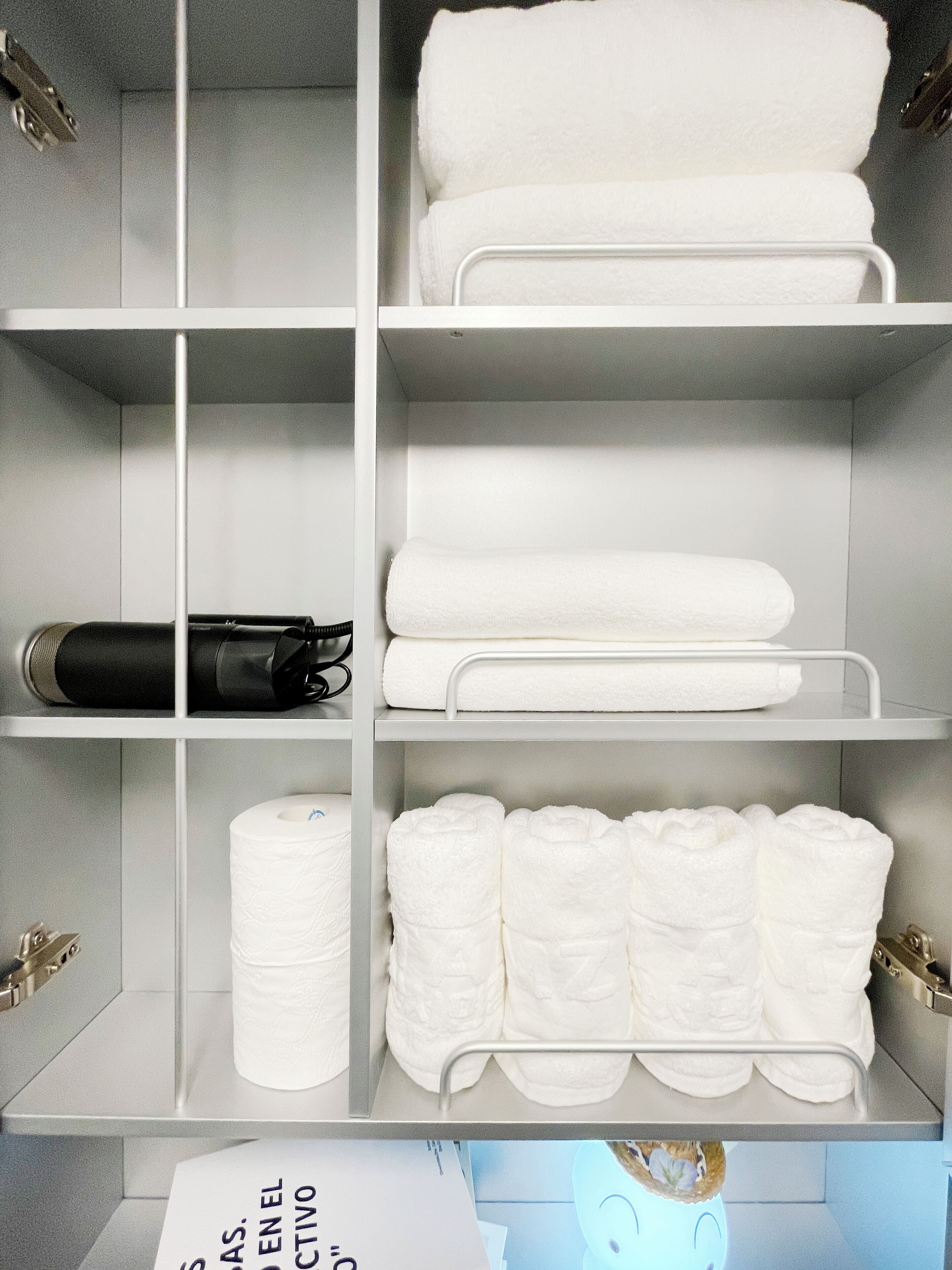 Un baño de hotel con repisas blancas que sostienen toallas, un secador de pelo negro y un rollo de papel higiénico.