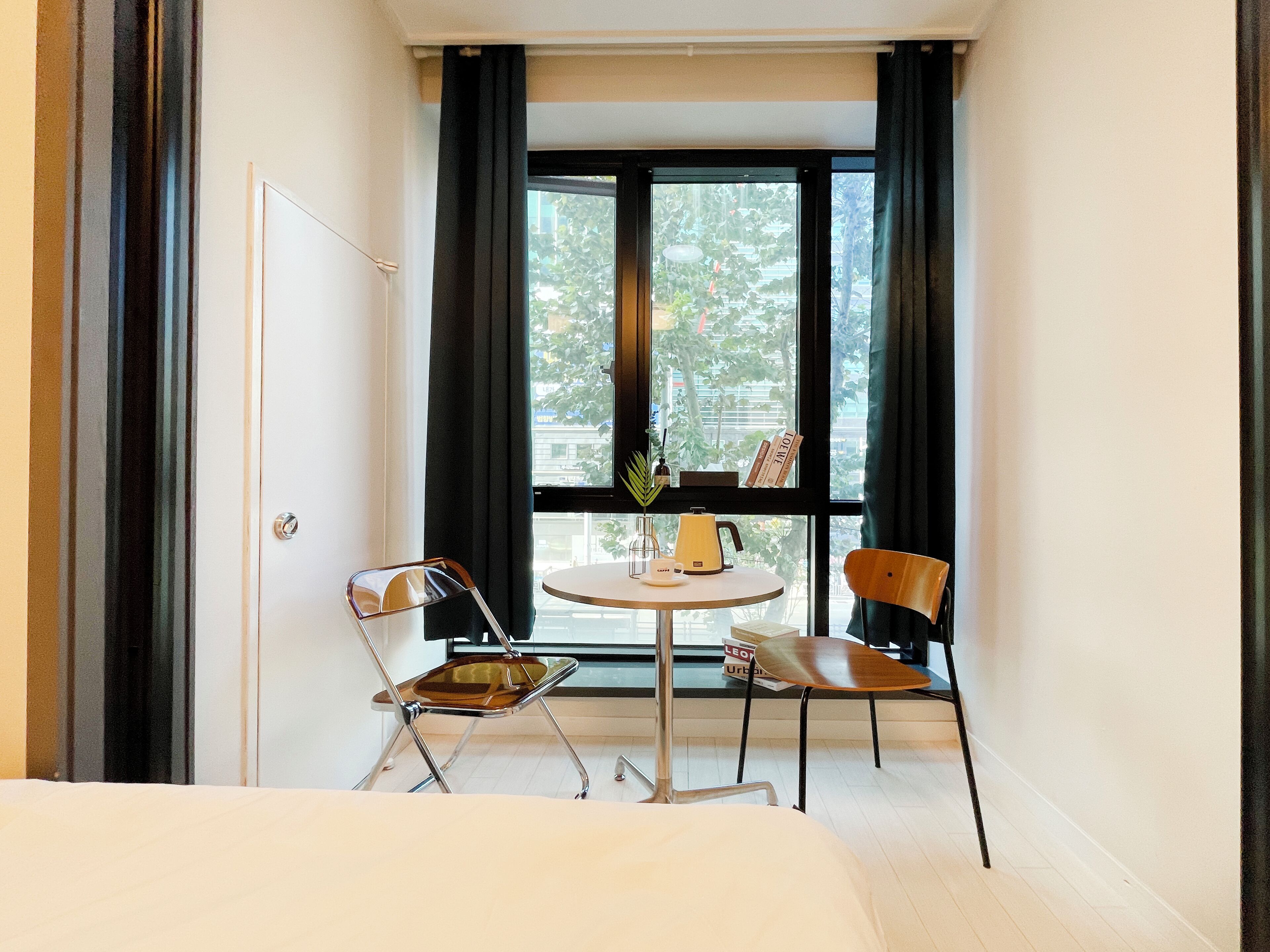 A hotel room with a bed, a small table with a kettle, two chairs, a window with curtains, and a door.