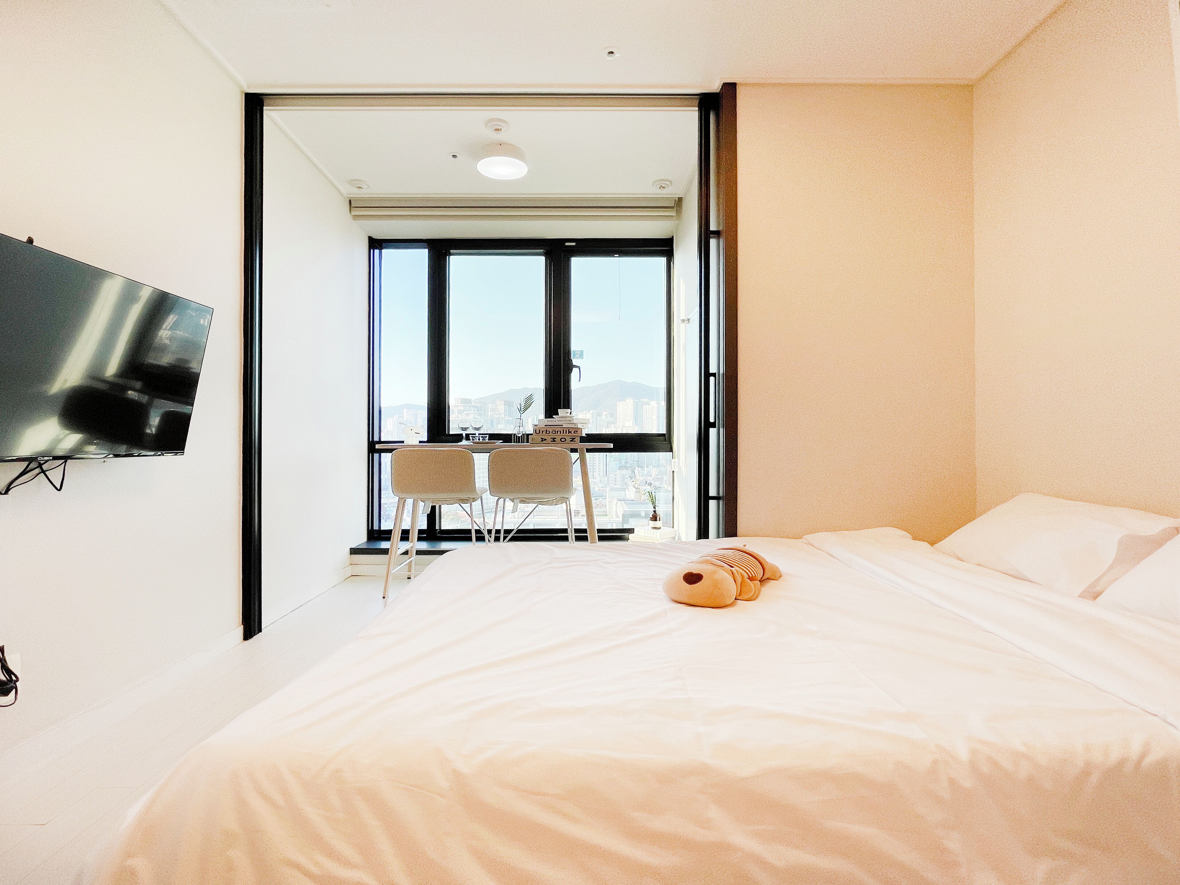 A modern hotel room with a large bed, a flat-screen TV mounted on the wall, a small table with chairs, and a view of the city through large windows.