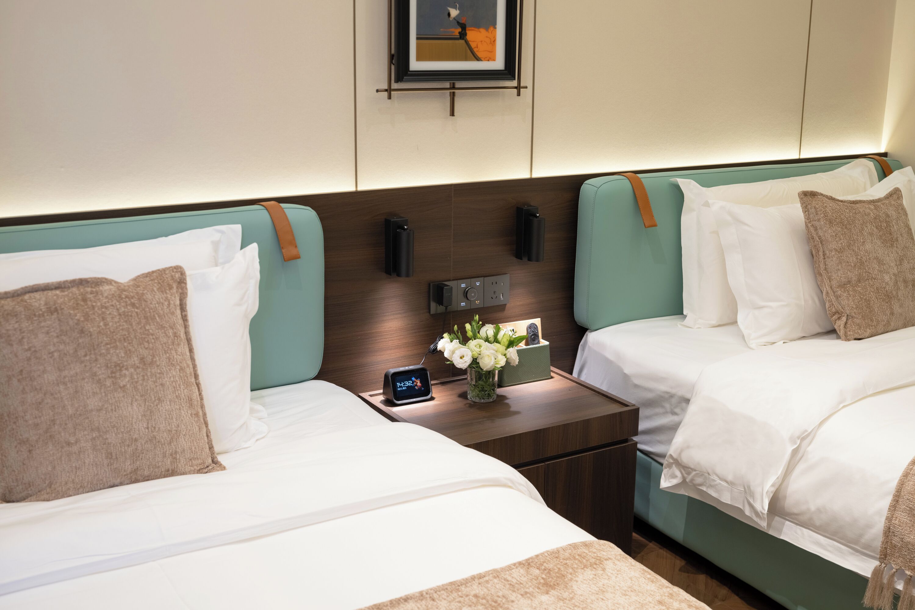 Habitación de hotel con dos camas, una mesita de noche con un reloj y un florero, y un cuadro en la pared.
