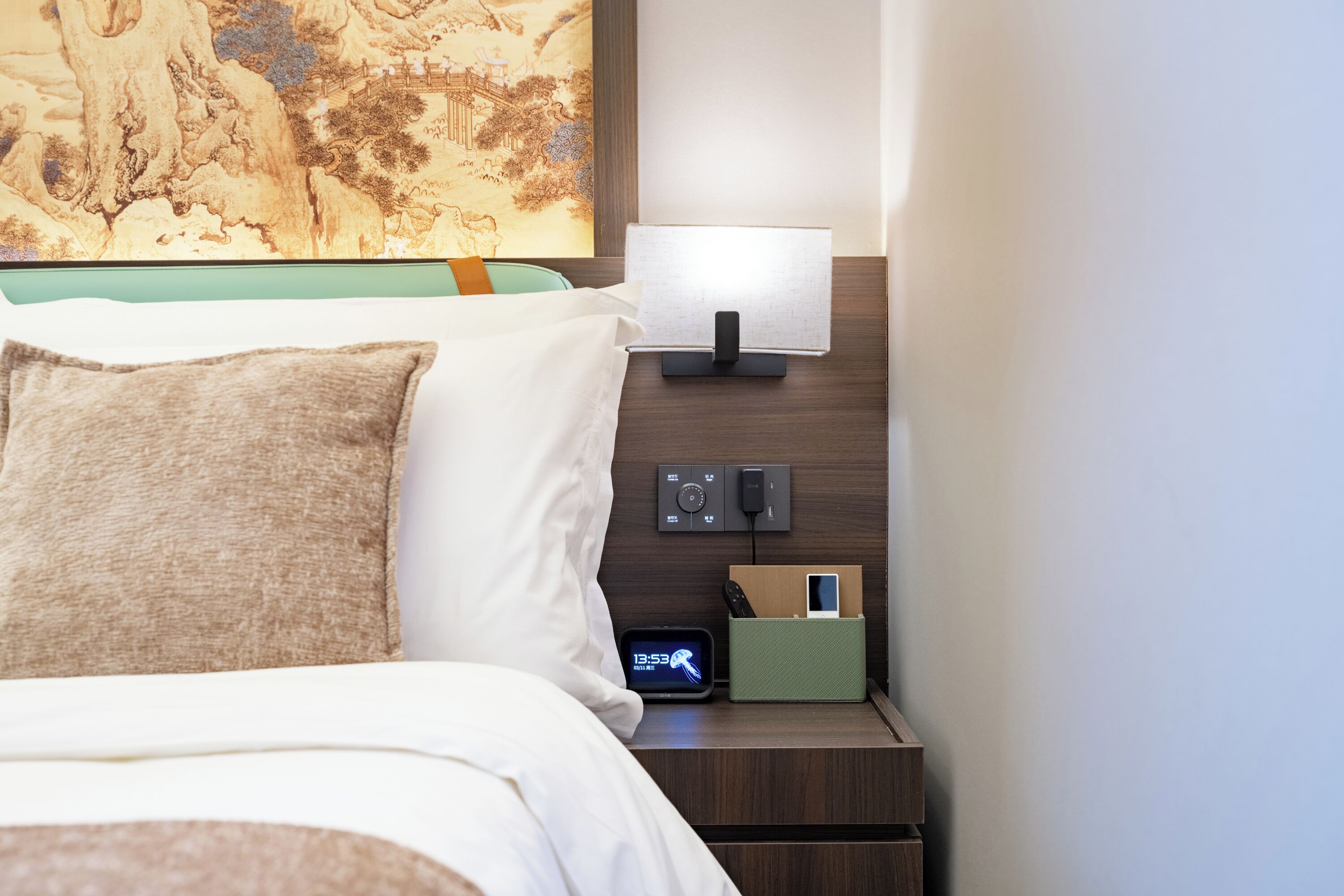A neatly made bed with pillows, a bedside table with a lamp, a clock, and a charging station.