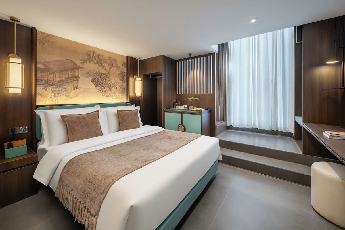 Ein modernes Hotelzimmer mit einem großen Bett, einem Schreibtisch, einem Sessel, einem Nachttisch, einem an der Wand befestigten Fernseher und einem großen Fenster mit Vorhängen.