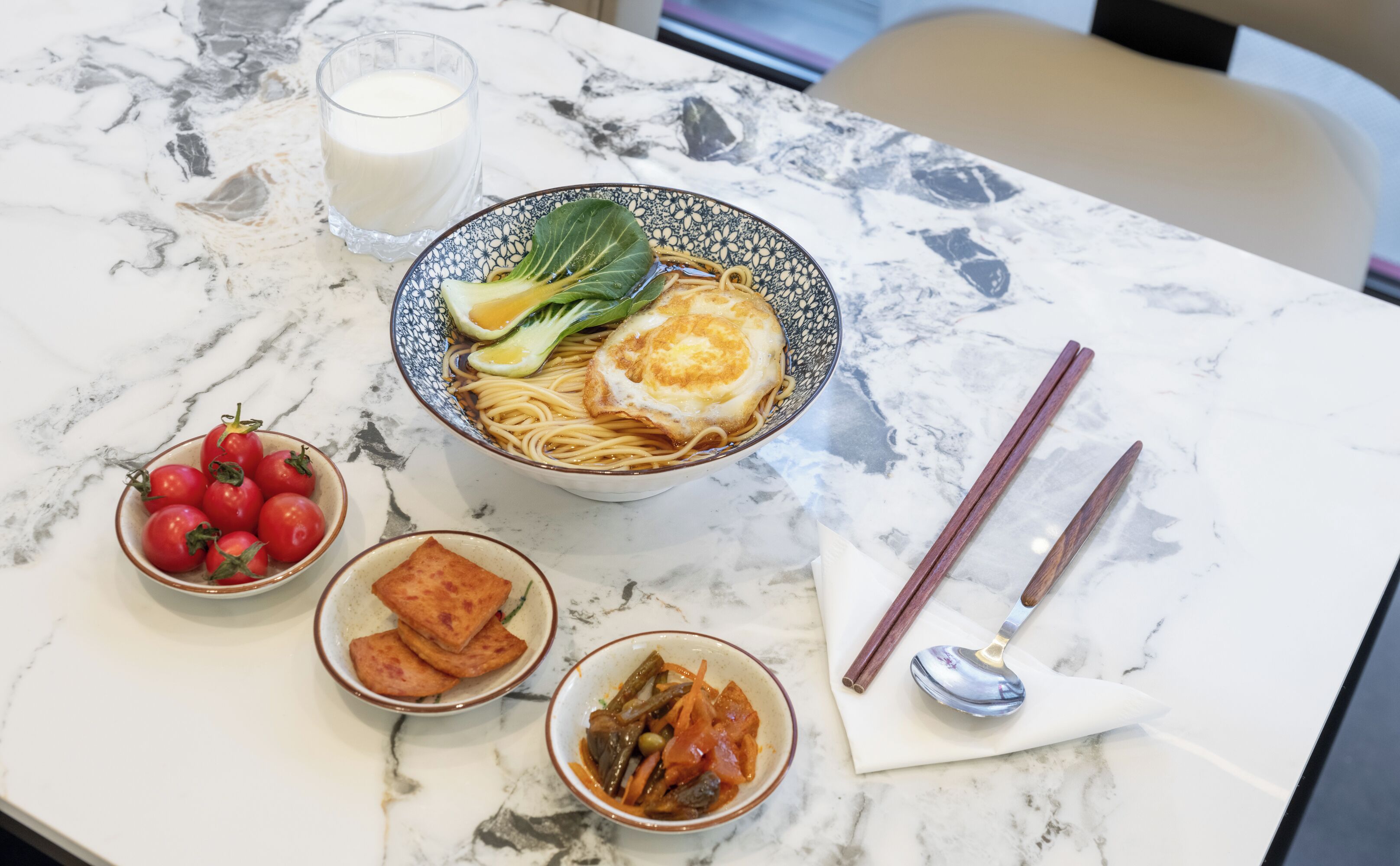 Una mesa con un tazón de fideos cubierto con un huevo frito, un vaso de leche, un tazón de tomates cherry, un tazón de tofu frito y una cuchara.