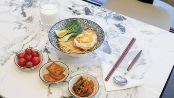 Una mesa con un tazón de fideos cubierto con un huevo frito, un vaso de leche, un tazón de tomates cherry, un tazón de tofu frito y una cuchara.