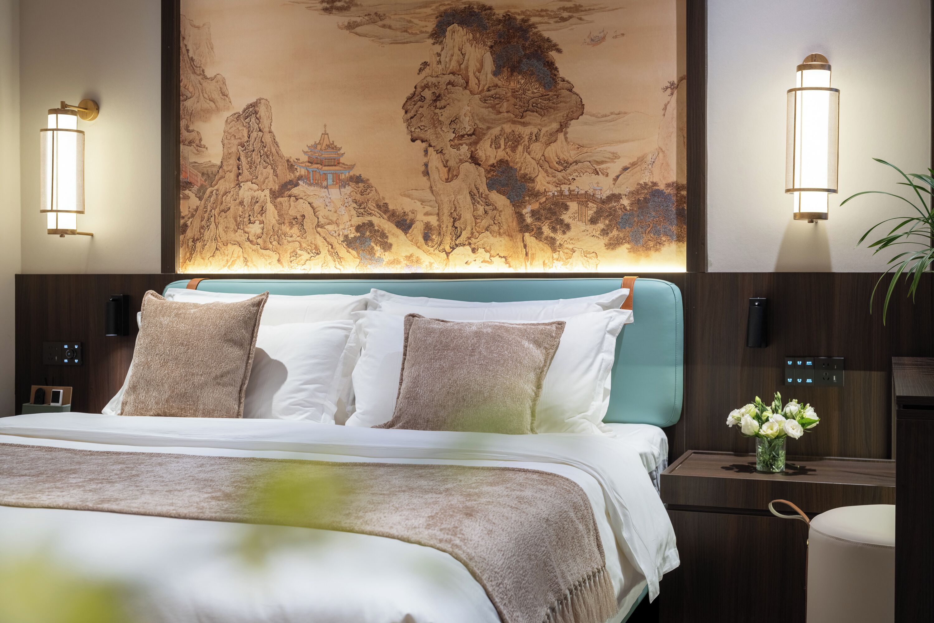 A hotel room with a bed, wall-mounted light fixtures, a large mural, and a nightstand with a vase of flowers.