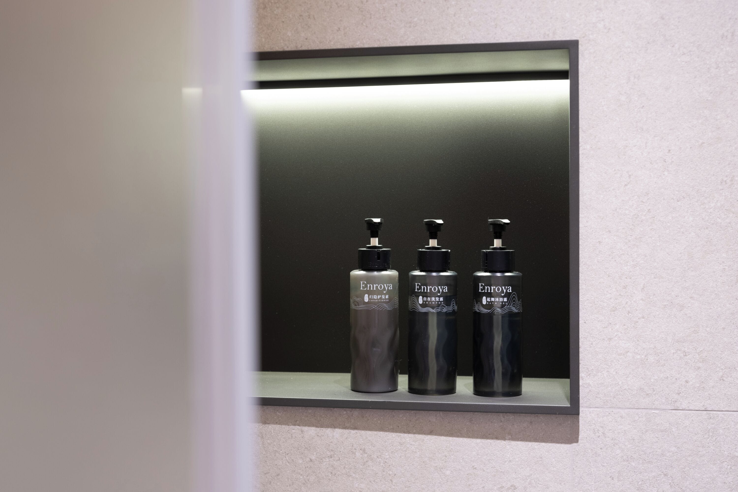Un baño con espejo, tres botellas de Enroya y una repisa iluminada.