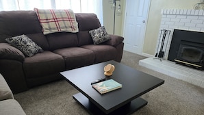 Un salon avec un canapé marron foncé, des coussins à motifs, une table basse noire sur laquelle se trouvent un livre et un objet en bois, et une cheminée dont le foyer est entouré de carreaux blancs.