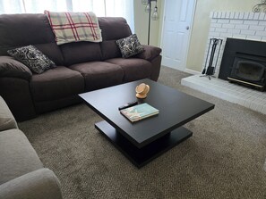 Ein Wohnzimmer mit einer dunkelbraunen Couch, gemusterten Kissen, einem schwarzen Couchtisch mit einem Buch und einem Holzobjekt sowie einem Kamin mit weiß gefliestem Rahmen.