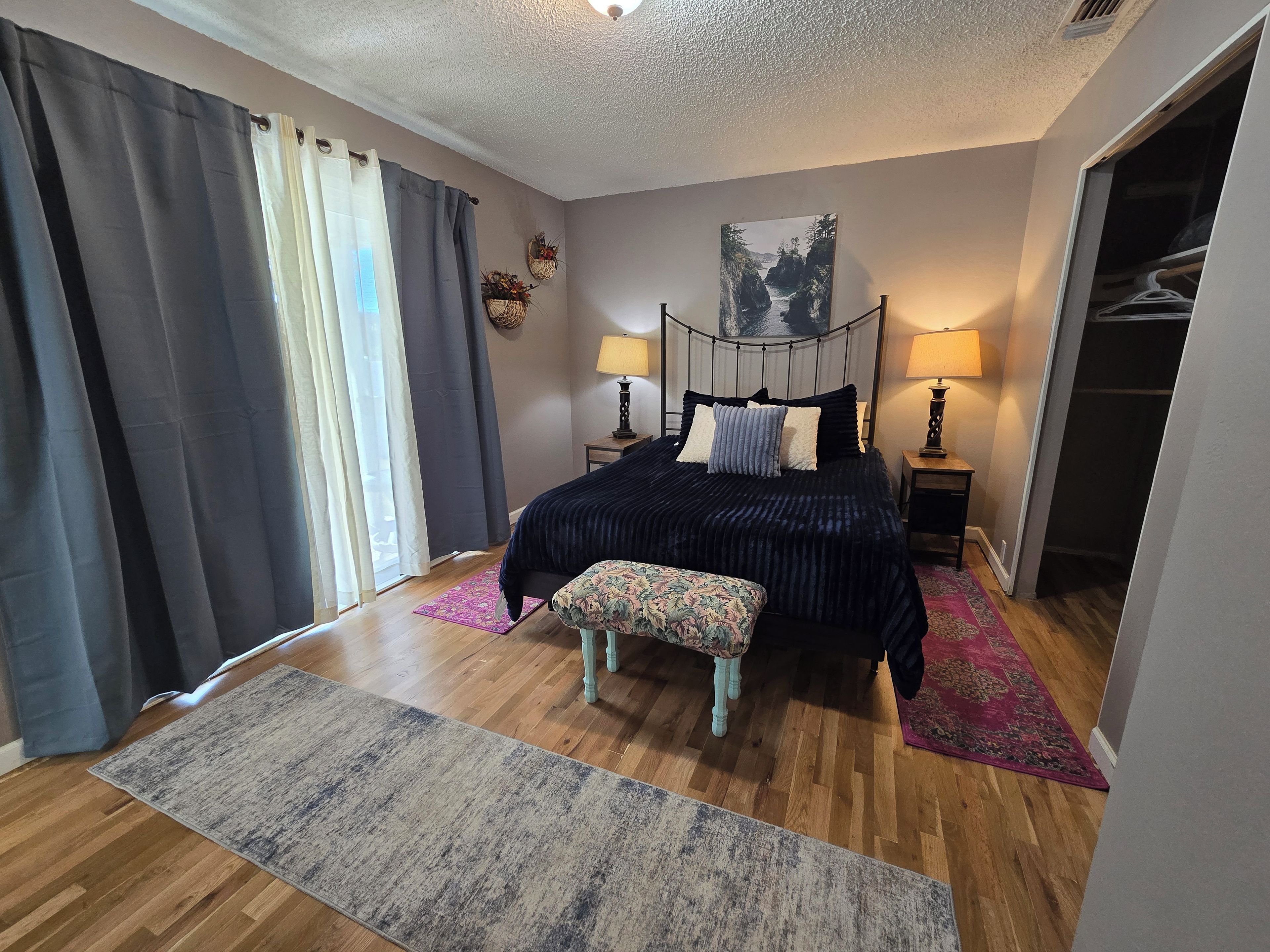 A bedroom with a bed, bedside lamps, a bench, a closet, and a view of a landscape painting.