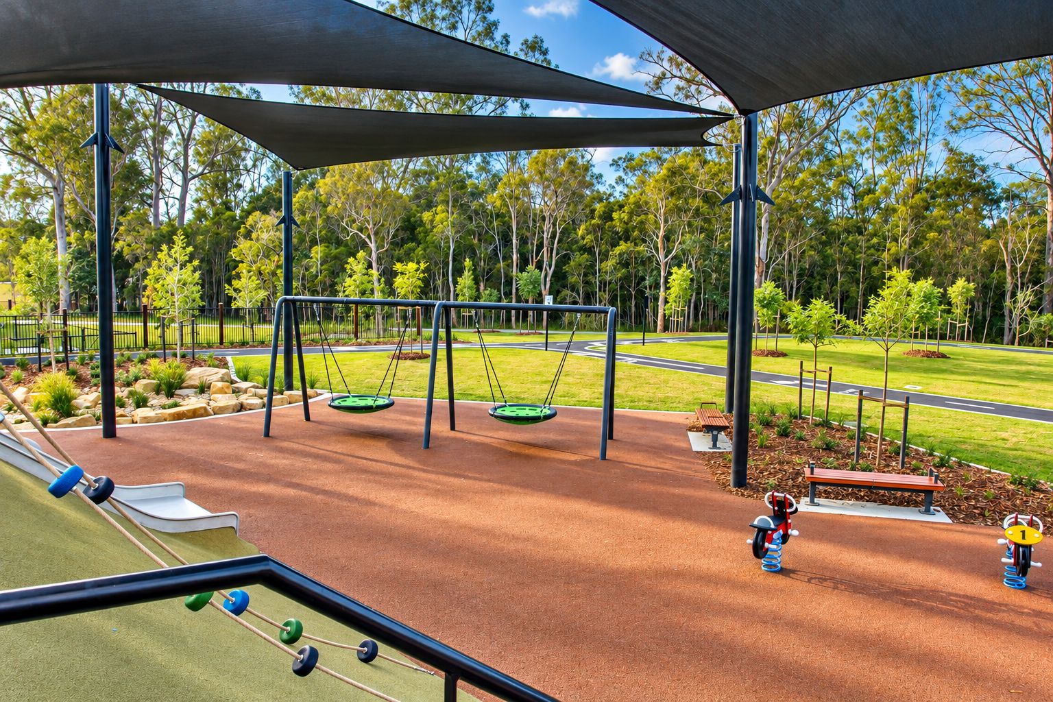A playground with swings and a slide, surrounded by trees and a road.