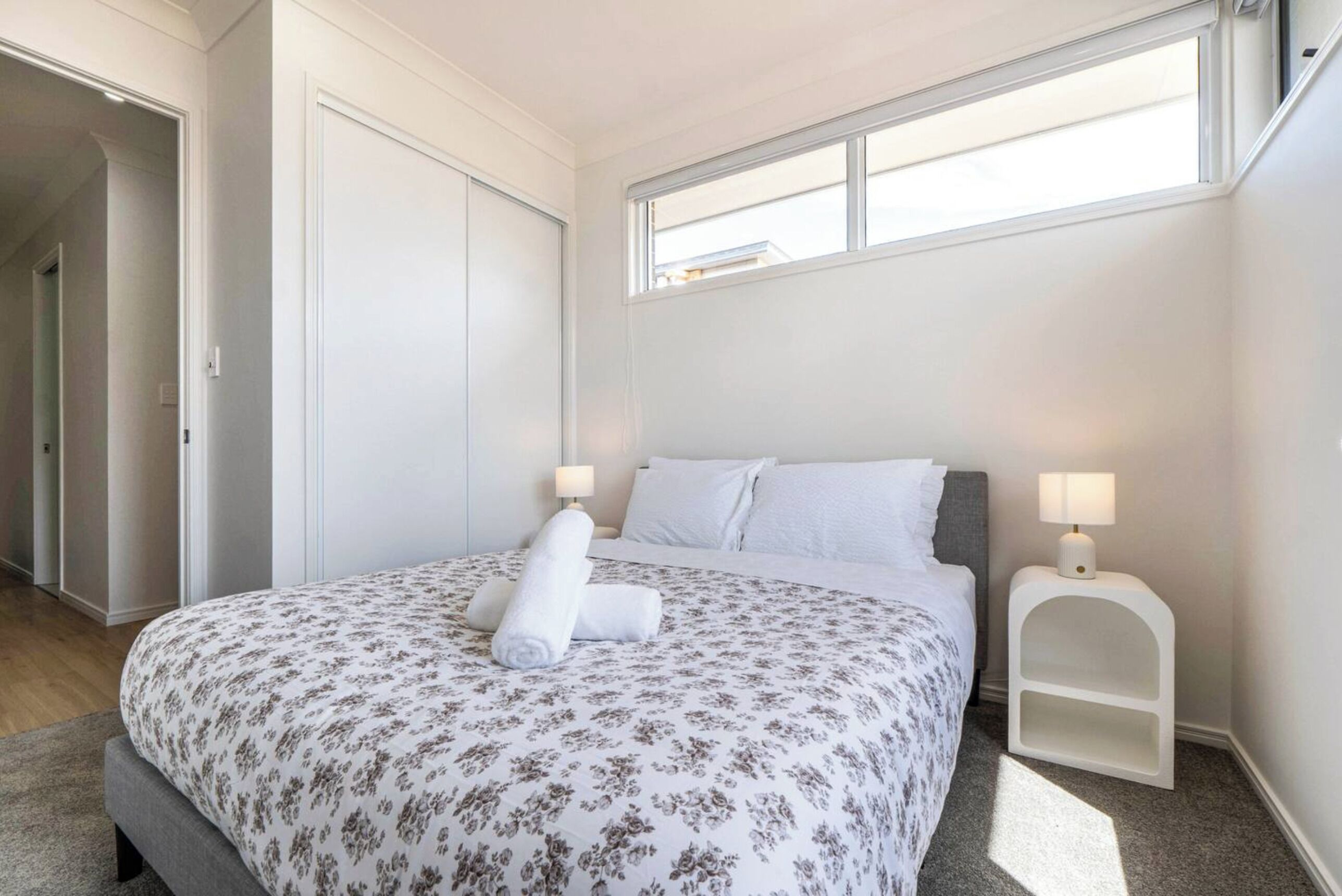 A neatly made bed with floral bedding, a bedside lamp, and a view of a hallway.